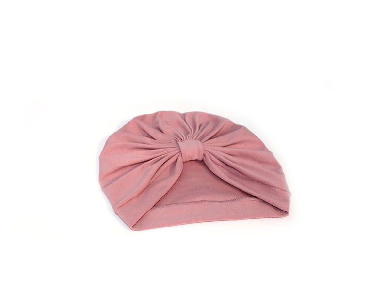 May include: A dusty rose turban-style hat. The hat is made of soft fabric and features a gathered design at the top, creating a stylish, knotted effect. The head covering is set against a white background.