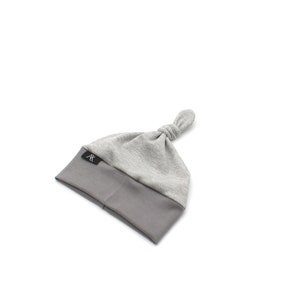 May include: A gray baby knot hat with a textured top and a solid gray brim. The hat has a knotted top and a small black label with white text. The hat is designed for infants.