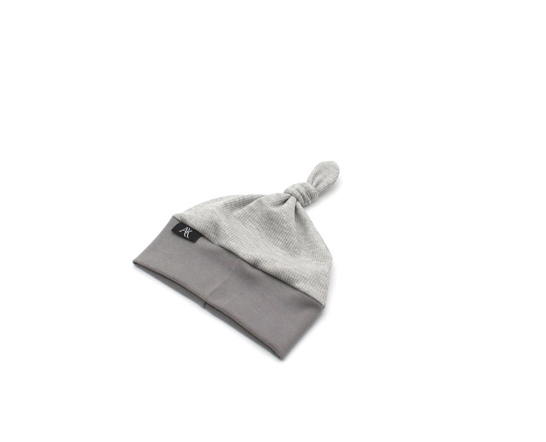 May include: A gray baby knot hat with a textured top and a solid gray brim. The hat has a knotted top and a small black label with white text. The hat is designed for infants.