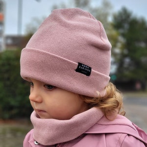 May include: A light pink beanie hat and matching neck warmer set. The beanie has a folded brim and a small black tag with white text that reads "ANNA KARINNA HANDMADE". The set is made of a soft, knitted material.