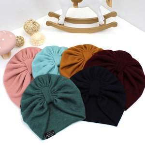 May include: A collection of seven colorful baby turbans in shades of pink, teal, mustard, burgundy, dark green, and navy blue. Each turban features a gathered design and a small label. A white rocking horse and decorative items are in the background.