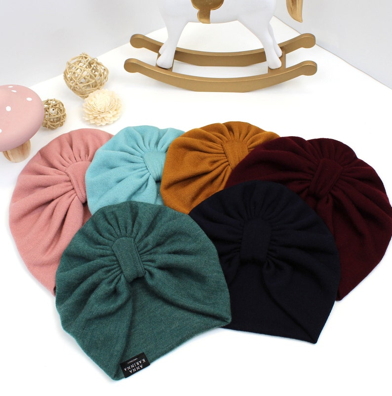 May include: A collection of seven colorful baby turbans in shades of pink, teal, mustard, burgundy, dark green, and navy blue. Each turban features a gathered design and a small label. A white rocking horse and decorative items are in the background.