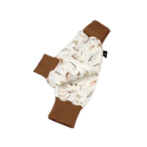 May include: Baby trousers featuring a cream-coloured fabric with a floral pattern in shades of brown and green. The cuffs are a solid brown colour. The trousers are made of soft material.