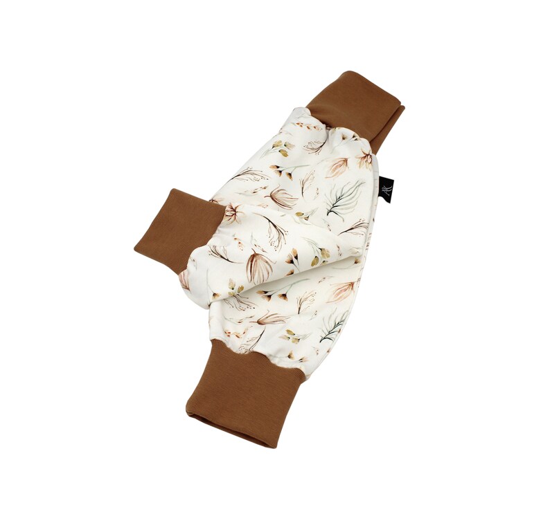 May include: Baby trousers featuring a cream-coloured fabric with a floral pattern in shades of brown and green. The cuffs are a solid brown colour. The trousers are made of soft material.