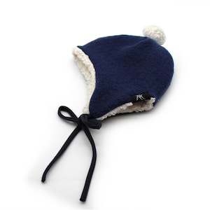 Peut inclure: Un bonnet d'hiver bleu marine avec une doublure en sherpa blanc et un pompon blanc sur le dessus. Le bonnet pr&eacute;sente des oreillettes et deux longs liens en tissu bleu marine pour attacher sous le menton. Une petite &eacute;tiquette noire avec les lettres blanches AK est fix&eacute;e.