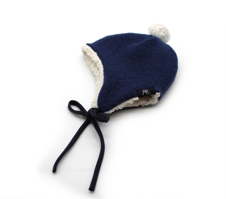 Peut inclure: Un bonnet d'hiver bleu marine avec une doublure en sherpa blanc et un pompon blanc sur le dessus. Le bonnet pr&eacute;sente des oreillettes et deux longs liens en tissu bleu marine pour attacher sous le menton. Une petite &eacute;tiquette noire avec les lettres blanches AK est fix&eacute;e.