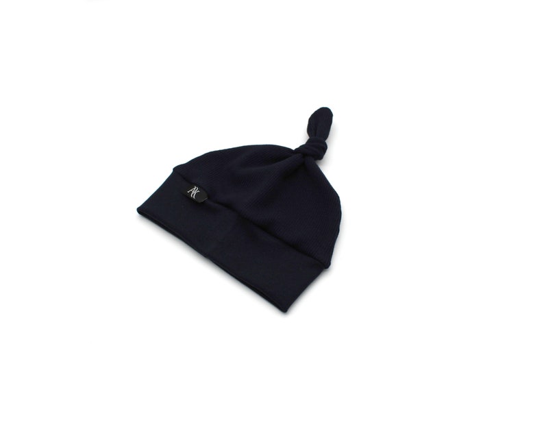 May include: A navy blue baby knot hat with a small, round, black logo on the folded brim. The hat is made of soft material and has a knotted top. The hat is designed for infants.