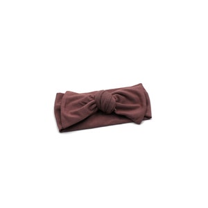 May include: A mauve-colored headband with a knotted bow detail. The fabric appears soft and stretchy, suitable for various head sizes. The headband is a solid color and rests on a white background.