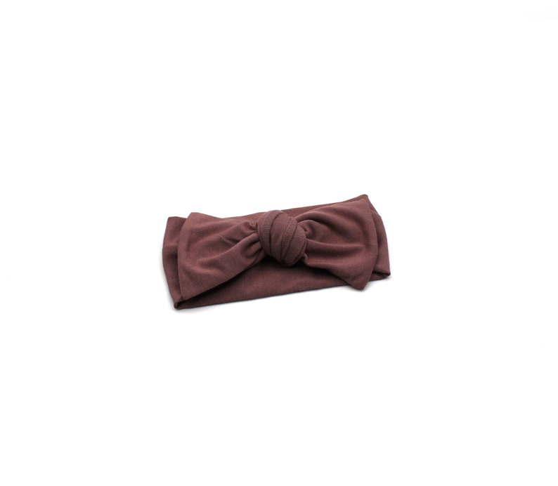 May include: A mauve-colored headband with a knotted bow detail. The fabric appears soft and stretchy, suitable for various head sizes. The headband is a solid color and rests on a white background.