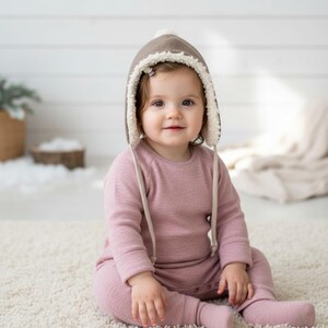 Peut inclure: Une grenouill&egrave;re &agrave; pieds rose poudr&eacute; &agrave; manches longues en tissu tricot&eacute; doux, dot&eacute;e d'une encolure ronde et de boutons-pression. La tenue est assortie d'un bonnet de style aviateur taupe doubl&eacute; de polaire sherpa blanche avec deux liens.