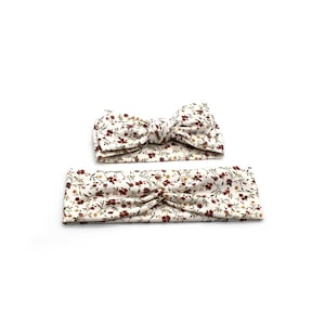 May include: Two floral headbands. One is a knotted bow, the other is a simple band. Both are white with a pattern of small red, yellow, and green flowers and leaves. The headbands are likely for babies or children.