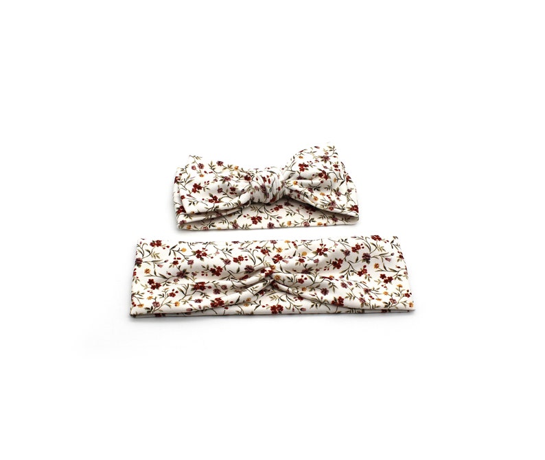 May include: Two floral headbands. One is a knotted bow, the other is a simple band. Both are white with a pattern of small red, yellow, and green flowers and leaves. The headbands are likely for babies or children.
