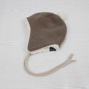 Peut inclure: Un bonnet &agrave; oreillettes marron avec une doublure en fausse peau de mouton blanche et un petit pompon blanc sur le dessus. Le bonnet pr&eacute;sente de longues lani&egrave;res beiges avec des extr&eacute;mit&eacute;s nou&eacute;es et une &eacute;tiquette noire avec ANNA MARIN.