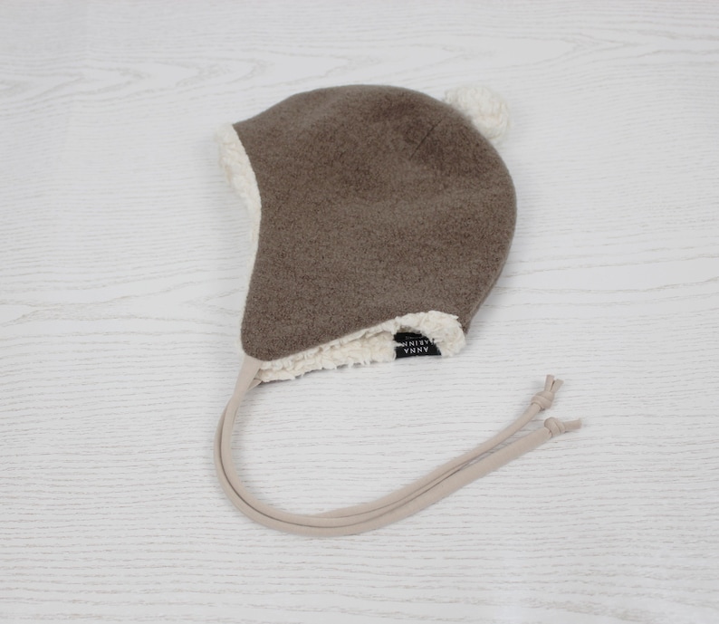 Peut inclure: Un bonnet &agrave; oreillettes marron avec une doublure en fausse peau de mouton blanche et un petit pompon blanc sur le dessus. Le bonnet pr&eacute;sente de longues lani&egrave;res beiges avec des extr&eacute;mit&eacute;s nou&eacute;es et une &eacute;tiquette noire avec ANNA MARIN.