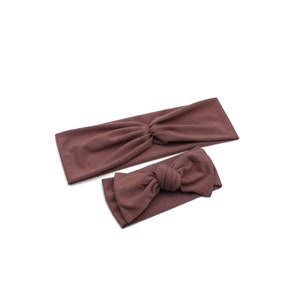 May include: Two mauve-colored headbands. One is a wide, twisted headband, and the other is a smaller headband with a bow. Both are made of a soft, solid-colored fabric. The headbands are likely for fashion or hair accessories.