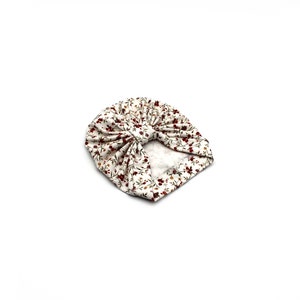 May include: A white turban-style baby hat with a floral pattern. The fabric features small, dark red flowers and green leaves scattered across a cream-colored background. The hat is knotted at the top.