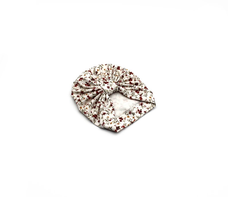 May include: A white turban-style baby hat with a floral pattern. The fabric features small, dark red flowers and green leaves scattered across a cream-colored background. The hat is knotted at the top.
