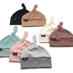 Peut inclure: Collection de sept bonnets pour bébés en tricot côtelé de différentes couleurs : marine, beige, crème, mauve, turquoise, rose et gris. Chaque bonnet a un nœud sur le dessus et une petite étiquette noire avec du texte blanc.