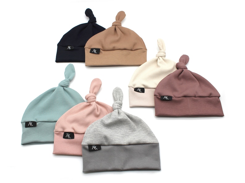 May include: A collection of seven ribbed knit baby hats in various colors, including navy, tan, cream, mauve, teal, pink, and gray. Each hat features a knotted top and a small black label with white text.
