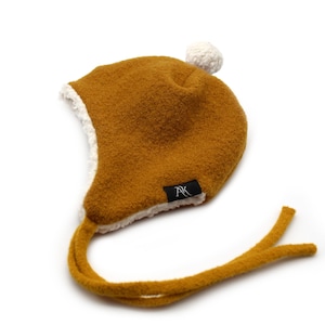 Peut inclure: Un bonnet &agrave; oreillettes jaune moutarde avec une doublure en polaire sherpa blanche et un pompon blanc sur le dessus. Le bonnet pr&eacute;sente de longs liens de menton jaune moutarde et une petite &eacute;tiquette noire avec les lettres AK en blanc.