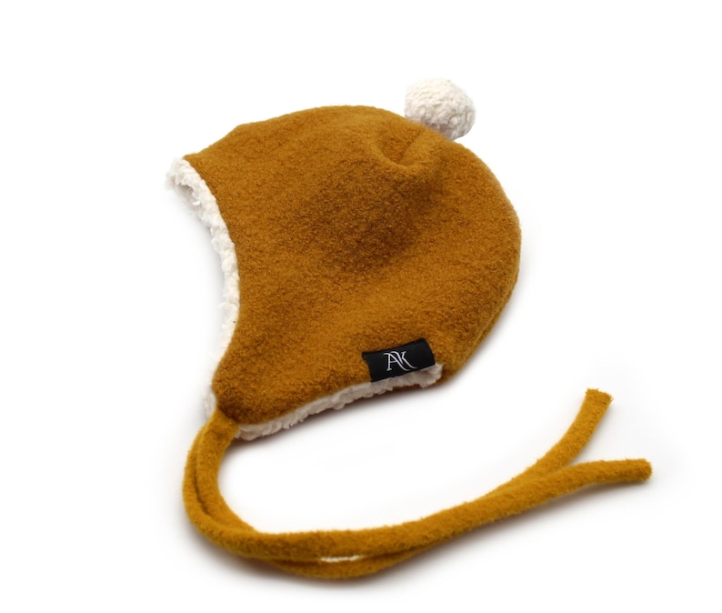 Peut inclure: Un bonnet &agrave; oreillettes jaune moutarde avec une doublure en polaire sherpa blanche et un pompon blanc sur le dessus. Le bonnet pr&eacute;sente de longs liens de menton jaune moutarde et une petite &eacute;tiquette noire avec les lettres AK en blanc.