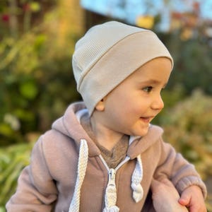 Może przedstawiać: Beżowa czapka z dzianiny żeberkowej. Czapkę nosi dziecko, kt&oacute;re ma r&oacute;wnież na sobie jasno brązową bluzę z kapturem z zamkiem błyskawicznym. Tło jest rozmyte, z zielonymi i brązowymi tonami.