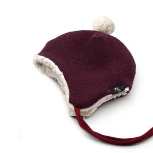 Peut inclure: Un bonnet d'hiver en feutre bordeaux fonc&eacute; avec une doublure en polaire sherpa cr&egrave;me et un pompon cr&egrave;me assorti sur le dessus. Le bonnet pr&eacute;sente des oreillettes, de longs liens en tissu bordeaux et une petite &eacute;tiquette noire avec un logo blanc.