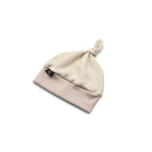 May include: A beige baby hat with a knotted top. The hat is two-toned, with a ribbed, light beige crown and a solid, darker beige brim. A small black tag is sewn on the brim. The hat is designed for infants.
