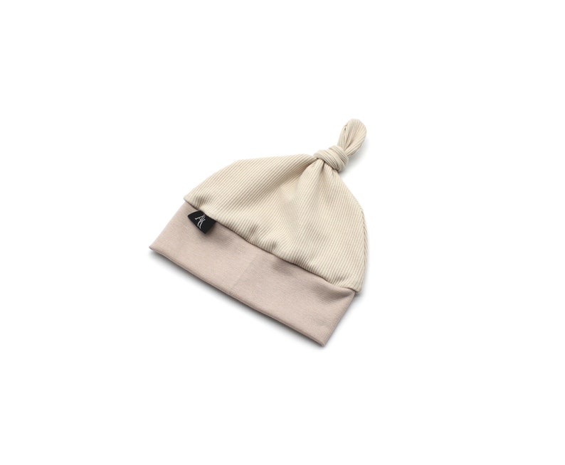 May include: A beige baby hat with a knotted top. The hat is two-toned, with a ribbed, light beige crown and a solid, darker beige brim. A small black tag is sewn on the brim. The hat is designed for infants.