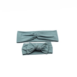 May include: Two teal headbands on a white background. One headband is a wide, twisted style. The other is a smaller headband with a bow. Both are made of a soft, stretchy fabric.
