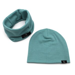 May include: A teal-colored beanie hat and neck gaiter set. Both items have a small black tag with white text. The beanie is a simple, close-fitting design. The neck gaiter is a tube shape, designed to be worn around the neck.