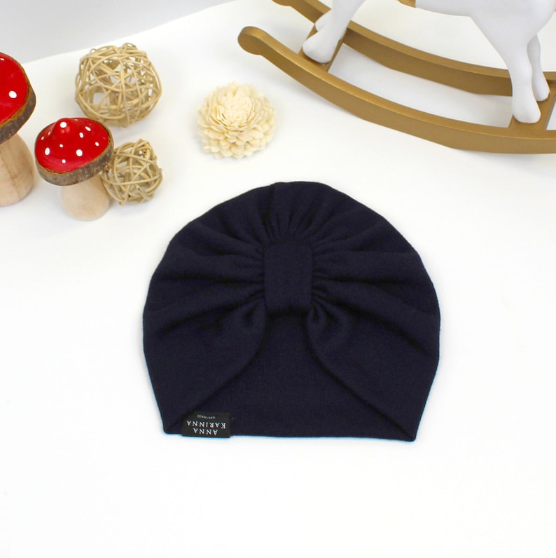 May include: Navy blue turban-style baby hat with a bow detail. The hat has a small rectangular tag with the text "ANNA KARINA" on it. The hat is on a white surface with decorative items in the background.