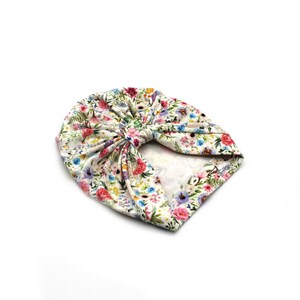 May include: A floral print turban in a cream color. The turban features a vibrant pattern of pink, red, blue, and yellow flowers with green leaves. The fabric is gathered and knotted at the top, creating a stylish head covering.