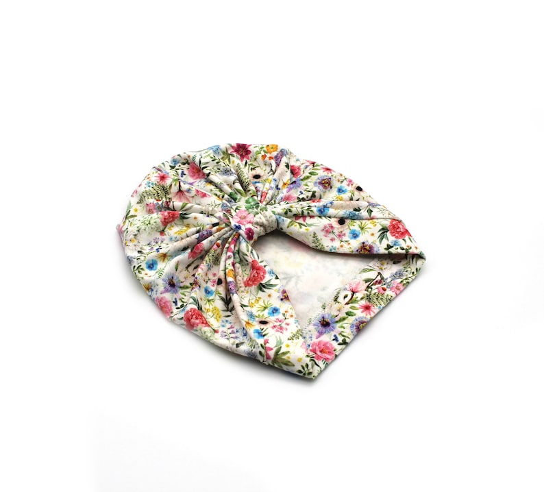 May include: A floral print turban in a cream color. The turban features a vibrant pattern of pink, red, blue, and yellow flowers with green leaves. The fabric is gathered and knotted at the top, creating a stylish head covering.