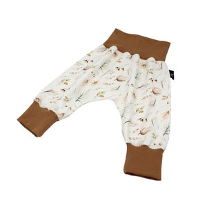 May include: Baby trousers with a white background and a pattern of brown and green leaves and feathers. The cuffs and waistband are solid brown. The trousers have a relaxed fit.