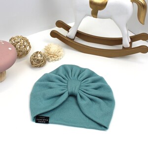 May include: A teal-colored baby turban hat with a large bow detail. The hat has a small black tag with white text that reads "ANNA KARINA". The hat is made of a soft, knitted material. Decorative items are in the background.