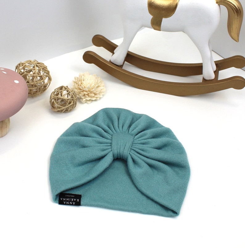 May include: A teal-colored baby turban hat with a large bow detail. The hat has a small black tag with white text that reads "ANNA KARINA". The hat is made of a soft, knitted material. Decorative items are in the background.