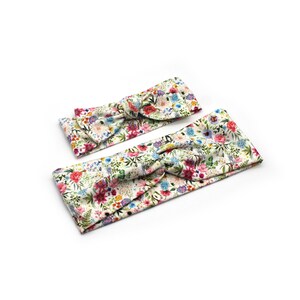May include: Two floral headbands with a knotted bow design. The headbands feature a white background with a colorful pattern of flowers in shades of pink, blue, yellow, and green. The headbands are likely for babies or children.