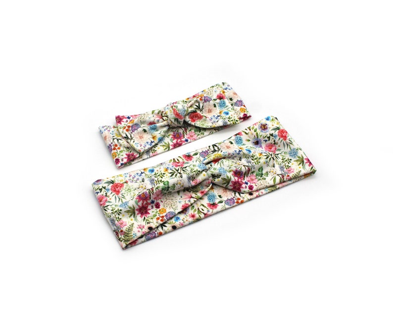 May include: Two floral headbands with a knotted bow design. The headbands feature a white background with a colorful pattern of flowers in shades of pink, blue, yellow, and green. The headbands are likely for babies or children.