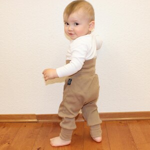 Könnte beinhalten: Ein Baby in einem weißen Langarmshirt und einer braunen Hose mit hohem Bund. Das Baby steht auf einem Holzboden und schaut über die Schulter. Die Hose hat ein kleines Etikett. Geeignet für Kleinkinder.