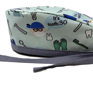May include: A light blue dental scrub cap with a gray brim and ties. The cap features a pattern of dental tools, teeth, and a cartoon dentist with the text "It's tooth:30".