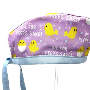 May include: Purple scrub hat with a repeating pattern of yellow chicks and the text "Oh, for Peep's Sake!" in white. The hat has a light blue tie.