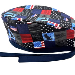 May include: A patterned scrub cap featuring a patchwork design with red, white, and blue American flag motifs, eagles, and the text "God Bless America, Land That I Love." The cap has a navy blue band and ties.