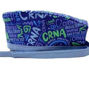 May include: A blue and green patterned scrub hat with a light blue tie. The hat has the text "CRNA" repeated in different styles and colors.