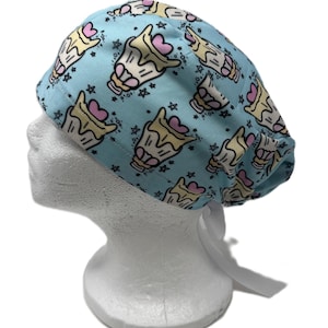 May include: A light blue scrub cap featuring a repeating pattern of stylized yellow and white dessert cups topped with pink hearts and small black stars. The cap has a rounded crown and includes white fabric ties at the back for an adjustable fit.