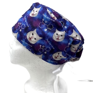 May include: A blue and purple scrub cap featuring a repeating pattern of white cat faces and chemical structure diagrams with the text Ketamine printed in white. The cap includes white fabric ties at the back for an adjustable fit.