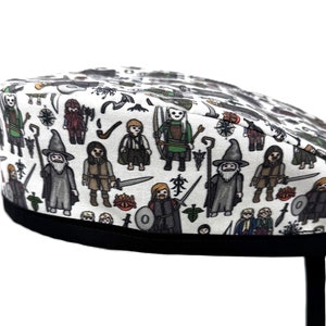 May include: White scrub hat with a black ribbon and tie. The hat is covered in a repeating pattern of cartoon characters from the Lord of the Rings.
