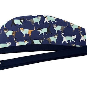 Może przedstawiać: Granatowy czepek chirurgiczny z powtarzającym się wzorem kotów w różnych kolorach, w tym białym, pomarańczowym i czarnym. Czepek ma ciemnoniebieski pasek i długie wiązania do mocowania. Nadruk z kotami jest na ciemnoniebieskim tle.