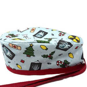 May include: A light blue surgical scrub cap with a red band and ties. The cap is patterned with Christmas-themed medical imagery, including x-rays, Christmas trees, and candy canes. The cap is designed for medical professionals.
