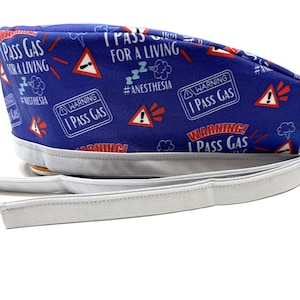 Op de afbeelding: Een blauwe chirurgische scrub cap met een witte voering en banden. De cap is bedrukt met de tekst "I PASS GAS FOR A LIVING", waarschuwingsborden en de hashtag "#ANESTHESIA".